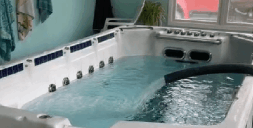 An overhead view of a white hot tub filled with turquoise water, featuring jets and control panels in an indoor bathroom or spa setting.