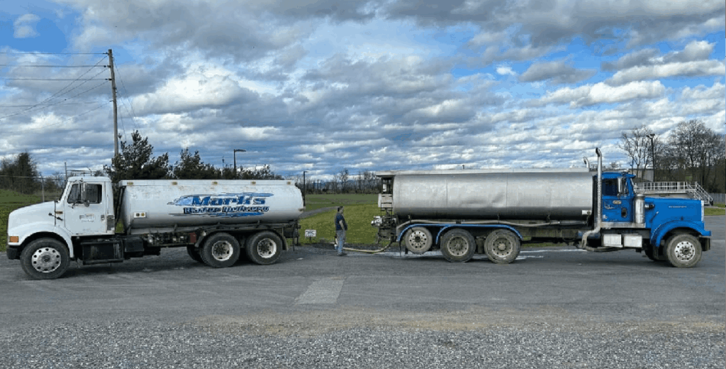 Two large tanker trucks are parked side by side on a rural roadway under a partly cloudy sky.