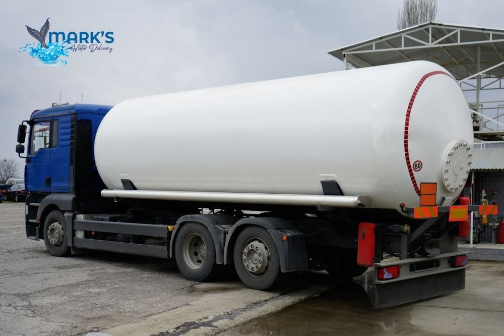 A large blue and white cylindrical tanker truck is positioned at an industrial facility with loading infrastructure visible in the background.